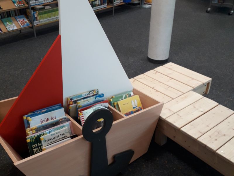 Ein Bilderbuchtrog in Form eines Schiffes mit vielen Bilderbüchern, daneben Bootsstege, im Hintergrund viele Bücherregale