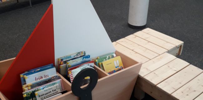 Ein Bilderbuchtrog in Form eines Schiffes mit vielen Bilderbüchern, daneben Bootsstege, im Hintergrund viele Bücherregale
